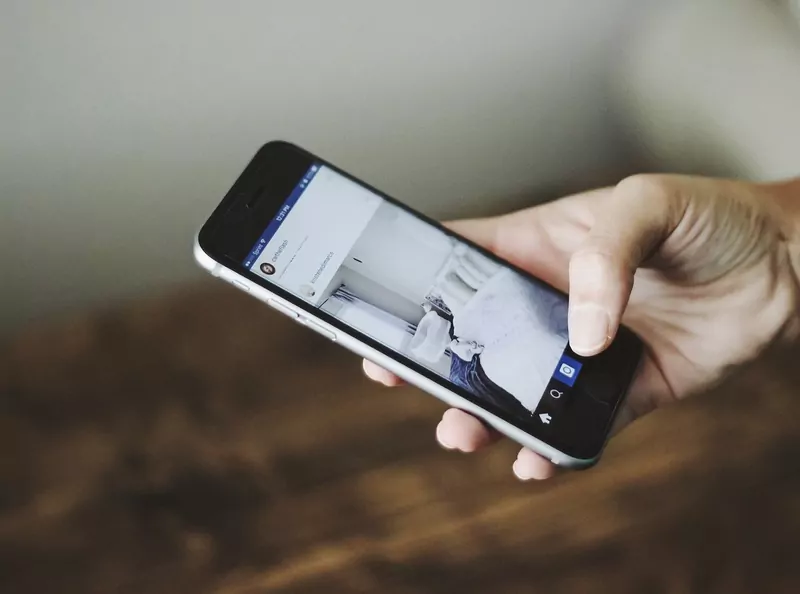 A hand holding a smartphone that shows an Instagram feed, in front of a wall and a wooden table