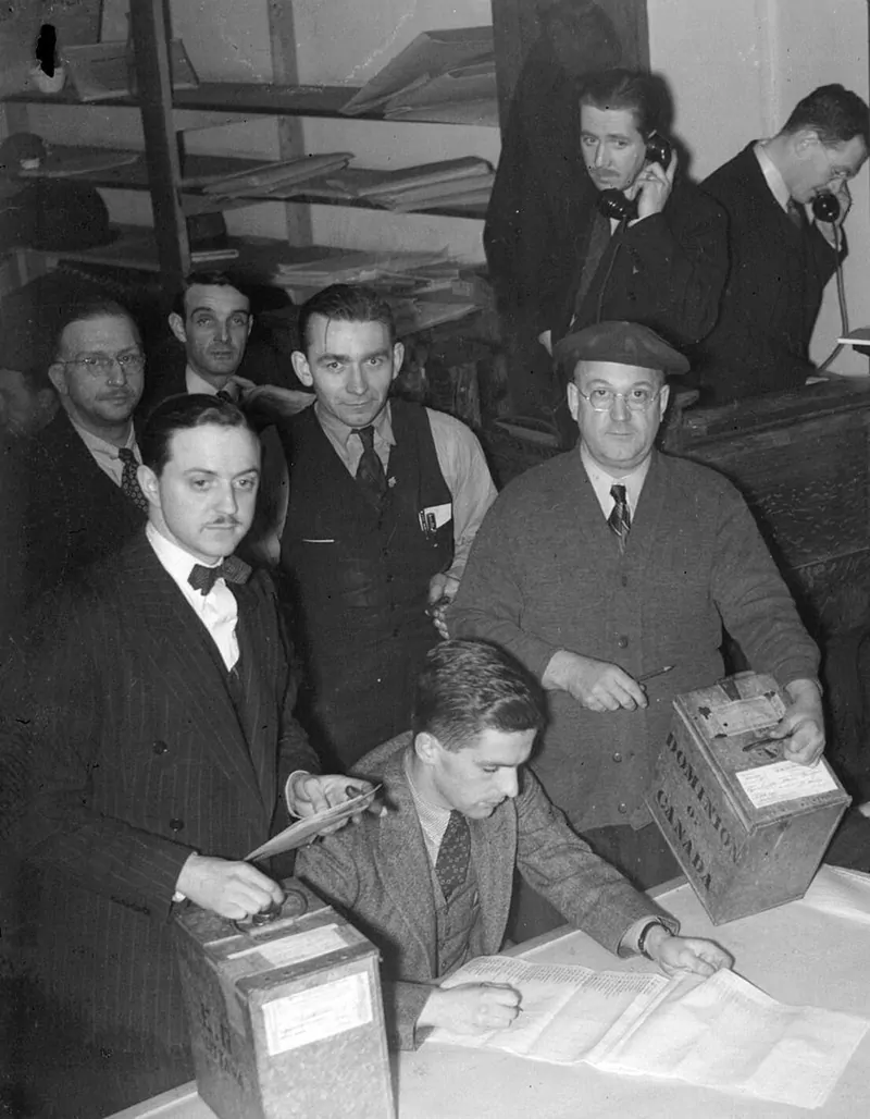 Black and white photo of election officials in suits counting paper ballots by hand at a table with wooden ballot boxes, two of them on telephones