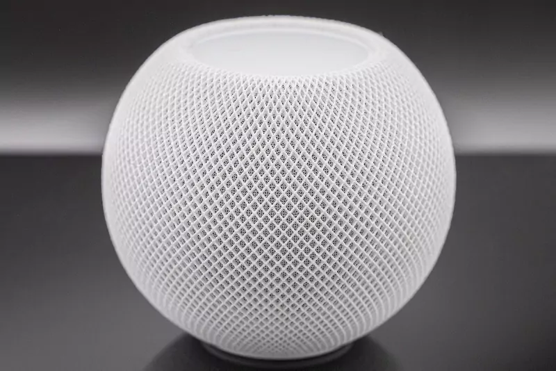 A white HomePod mini speaker covered in fine mesh fabric, shown close up against a dark glossy background