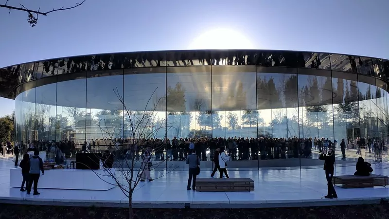 The round glass pavilion of the Steve Jobs Theater at Apple Park at sunset, with visitors walking on the white paving in front