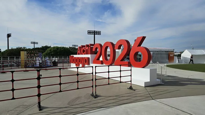 Giant red sign reading Aichi-Nagoya 2026 on a white plinth in a plaza beside Mizuho Park Athletic Stadium, with floodlight towers and trees behind it