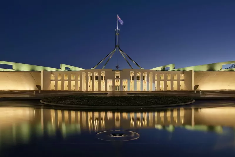 Parliament House in Canberra at dusk, with its tall flagpole above the lit building reflected in a pool