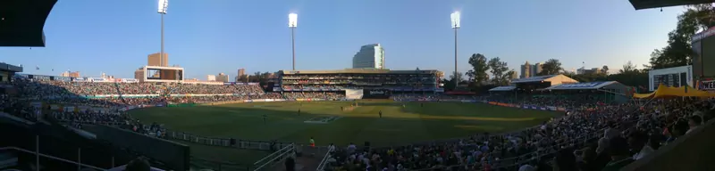 Panoramic view of Kingsmead cricket ground in Durban at dusk, with floodlights on, a packed crowd in the stands and players on the green outfield