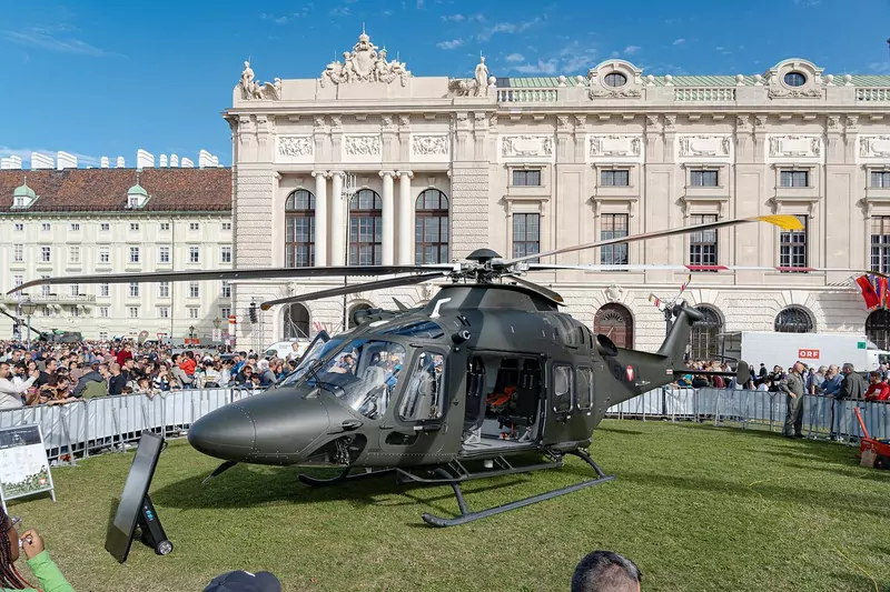 A dark green military helicopter on the grass of Heldenplatz in front of the Hofburg, with crowds behind barriers