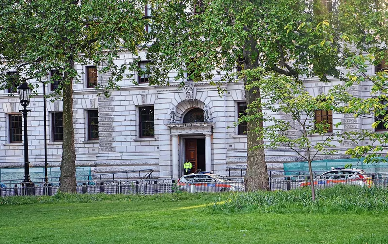 The pale stone HM Treasury building seen through trees across a lawn, with a police officer at the arched doorway and police cars outside