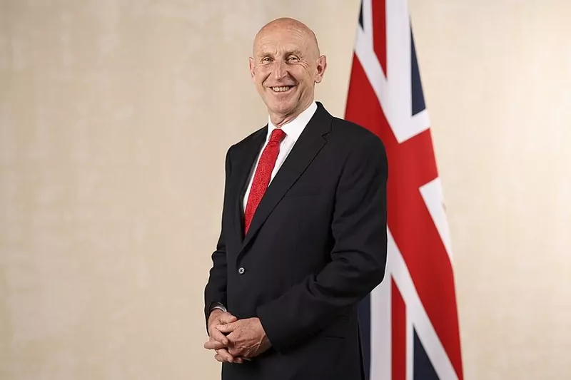John Healey in a dark suit and red tie, smiling, standing beside a Union flag