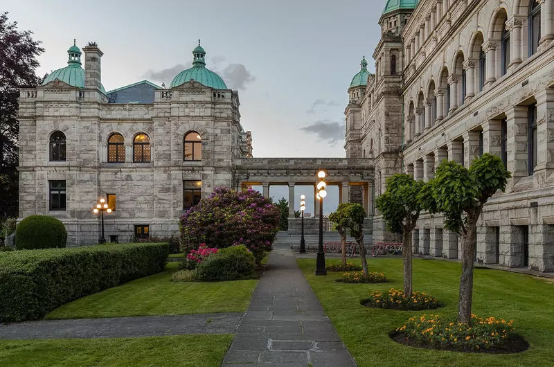The stone British Columbia Parliament Buildings in Victoria with green copper domes, seen from a garden path lined with trees and flowers
