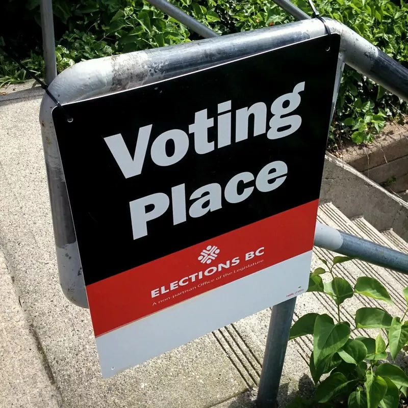 A black and red Elections BC sign reading Voting Place fixed to a metal railing beside steps and green bushes