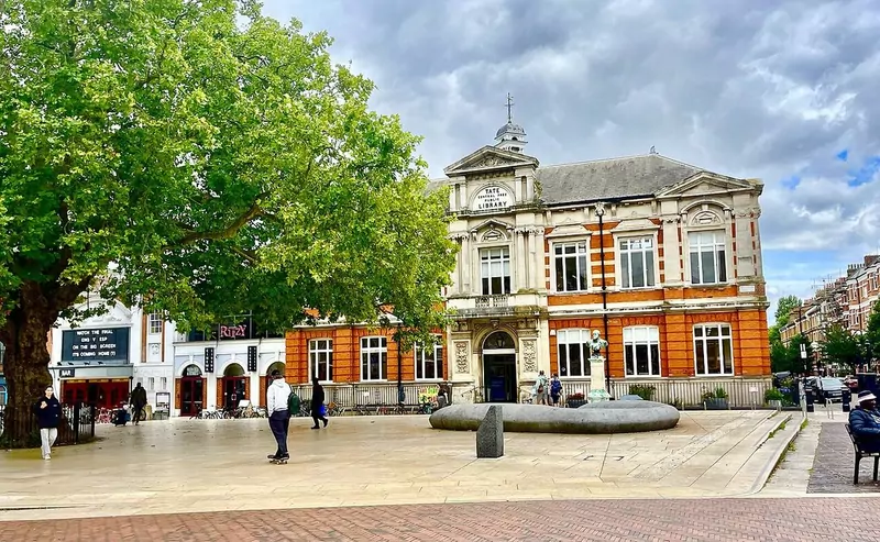 Brixton Library, a stone building with a clock tower, on a sunny square with trees