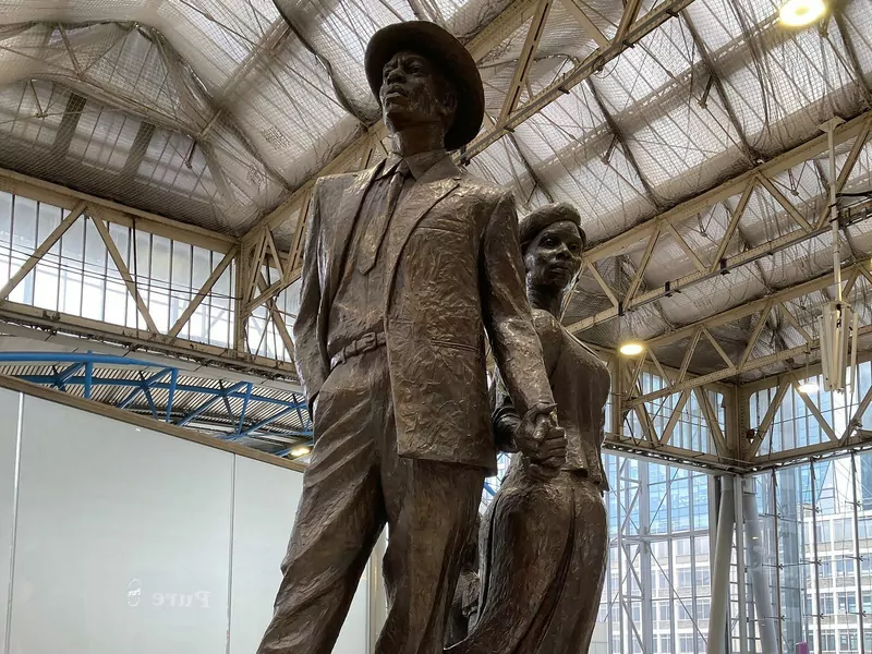 A bronze statue of a man in a suit and hat and a woman in a dress, holding hands, inside a station hall