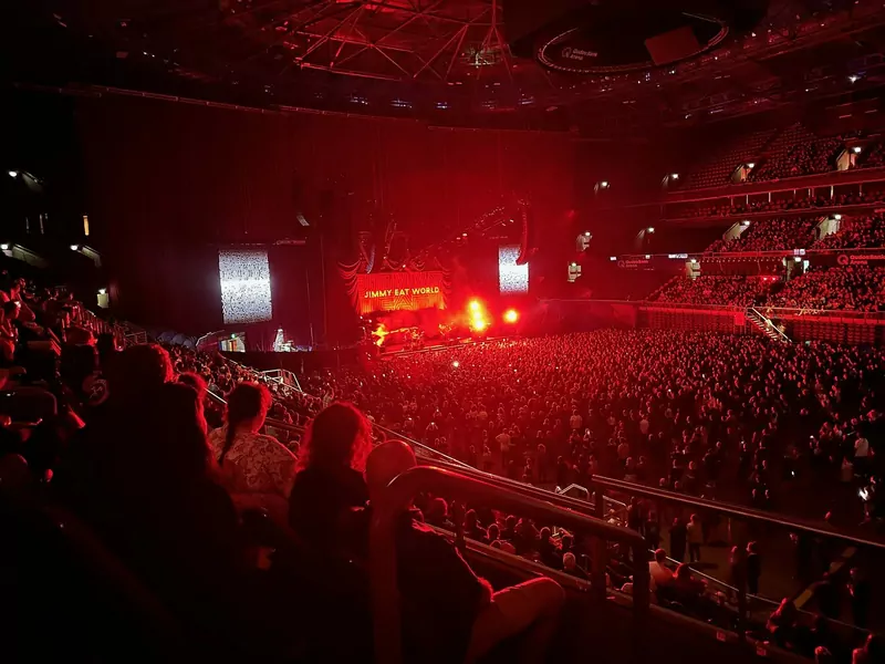 A packed arena crowd bathed in red light, facing a distant stage