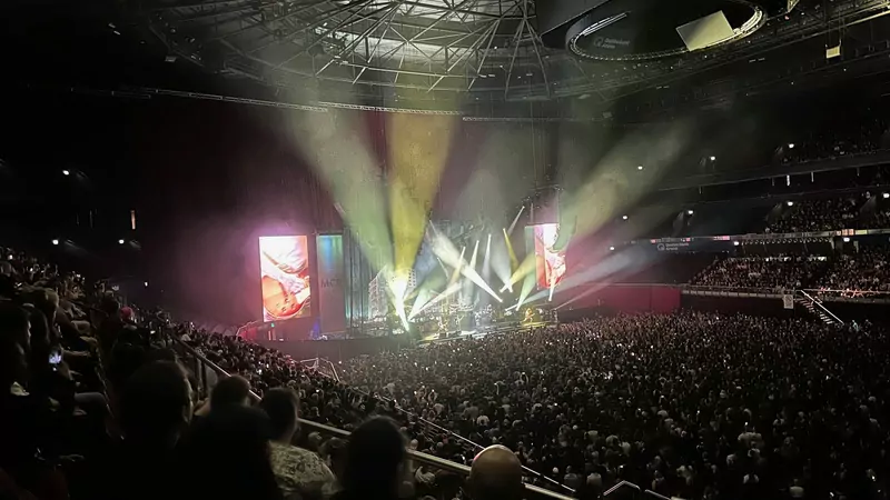 A large indoor arena with a lit stage and light beams in the distance and a packed crowd in the seats, during a My Chemical Romance concert