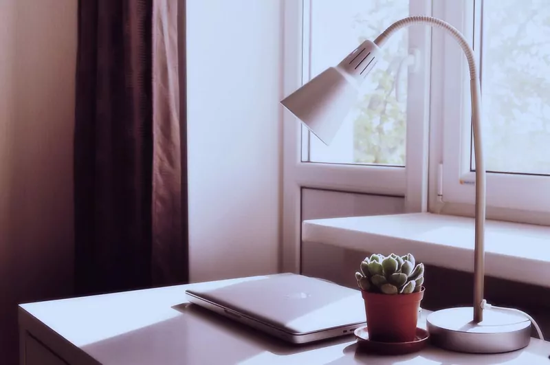 A closed laptop, a white desk lamp and a small succulent in a terracotta pot on a white desk by a window