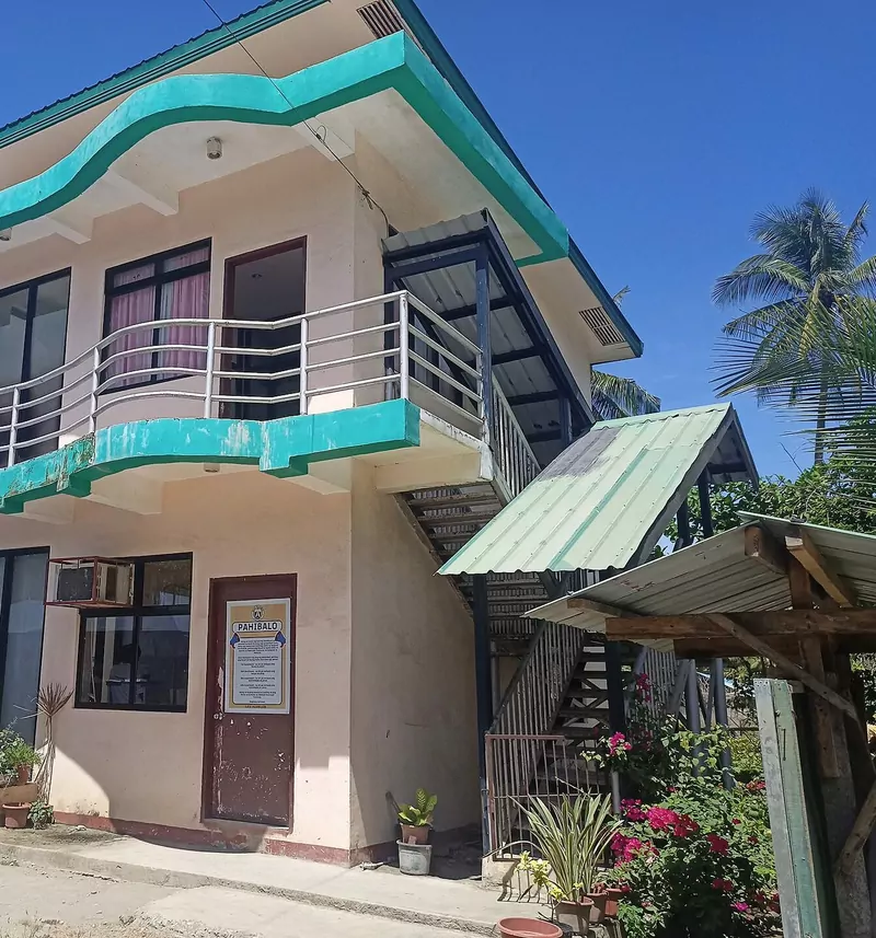 A cream two-storey barangay hall with a curved turquoise balcony, an outside staircase and flowering plants, under a blue sky