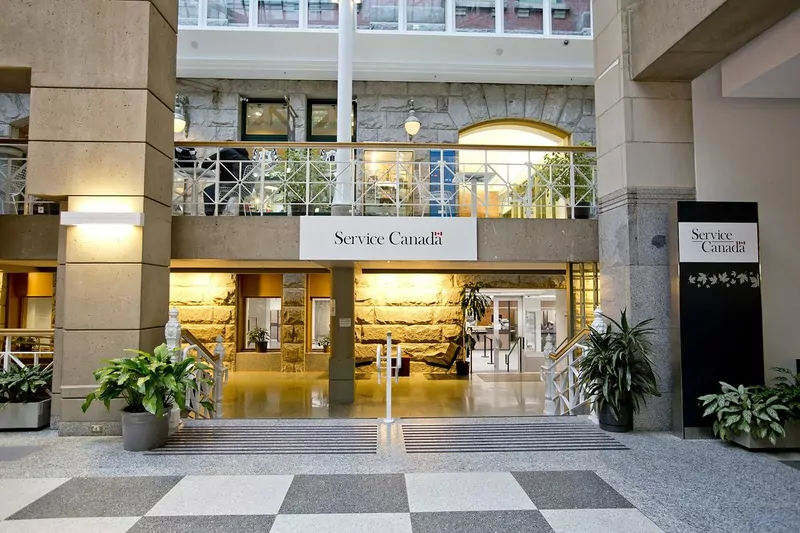 The entrance of a Service Canada centre inside a glass-roofed atrium with stone pillars, potted plants and a chequered floor