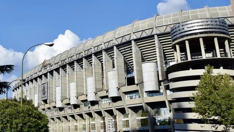 The long concrete outer wall of the Santiago Bernabéu stadium in Madrid under a blue sky, with the stadium name in letters on the roof and a round ramp tower at the right