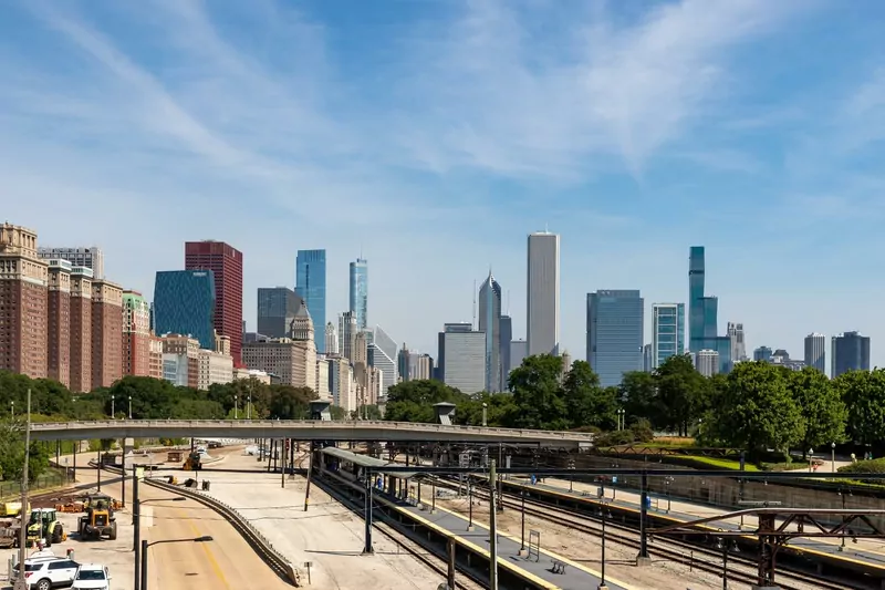 Chicago's downtown skyline on a sunny day seen from Grant Park, with tall buildings behind trees, a pedestrian bridge and railway tracks in the foreground
