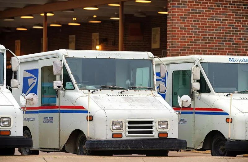 White US Postal Service delivery trucks parked in a row outside a brick post office