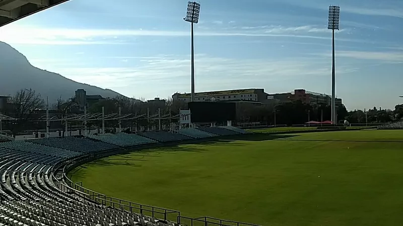 Newlands cricket ground in Cape Town in morning light, with rows of empty grey seats, a green outfield, two tall floodlight towers and a mountain behind