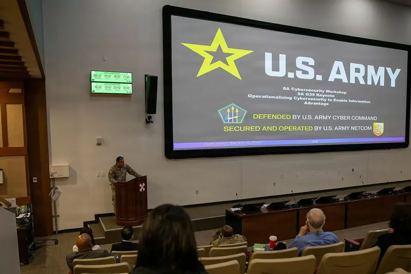 A speaker at a lectern in a lecture hall with a large screen showing a cybersecurity workshop title slide, with the audience in the foreground
