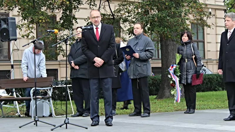 A man in a dark suit and red tie speaks at a microphone outdoors, with officials in coats standing behind him and a ribbon in the Czech colours on a wreath