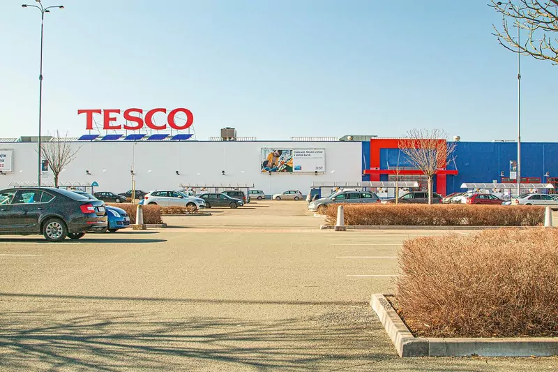 A large white Tesco hypermarket building with a red logo behind a mostly empty car park under a clear blue sky