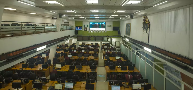 Rows of desks with computer screens on the trading floor of the Nigerian Stock Exchange in Lagos, with traders in maroon vests and a display board and Nigerian flag at the far end