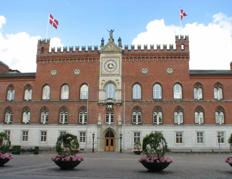 The red-brick town hall of Odense with a clock in its central tower, two Danish flags, a stone square and large flower planters in front