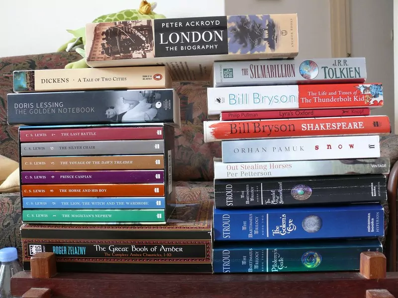 Two tall stacks of paperback novels on a sofa, including C. S. Lewis's Narnia books, Tolkien's The Silmarillion and books by Bill Bryson
