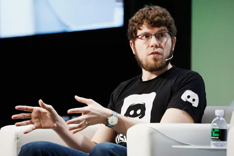 A bearded man in glasses and a black T-shirt with the Discord mascot, gesturing as he speaks on a stage