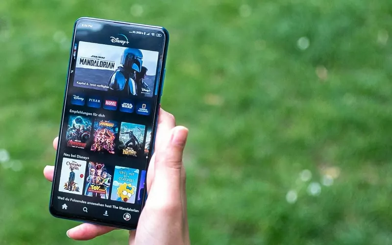 A hand holding a phone that shows the Disney+ app with a row of film posters, in front of green grass