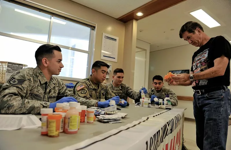 A man hands a prescription bottle to uniformed staff in blue gloves sorting collected medicines at a long table