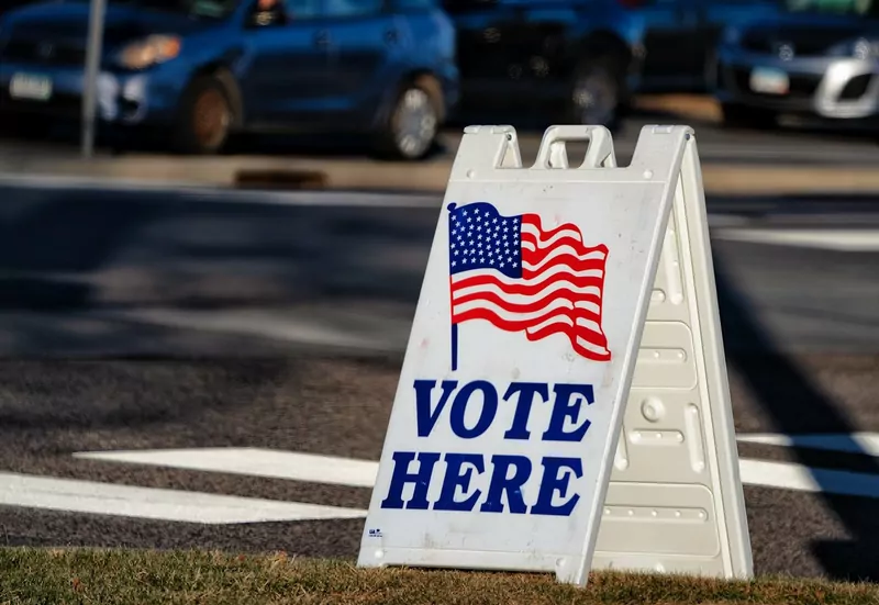 A white folding sign with an American flag and the words Vote Here standing on a sidewalk, with parked cars behind it