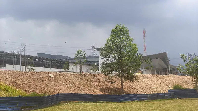 A new grey and white station building with a sloping roof behind a bank of bare earth and a young tree, under a dark grey sky, with a blue construction fence along the bottom edge