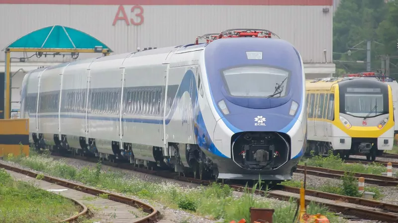 A streamlined blue, white and silver electric passenger train with the ECRL logo on its nose stands on a track in a depot beside a yellow metro train, in front of a large grey building marked A3, with green trees behind