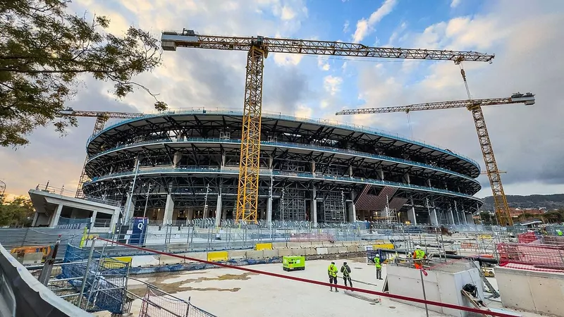 The steel frame of a large oval stadium with cranes above it and construction workers in front