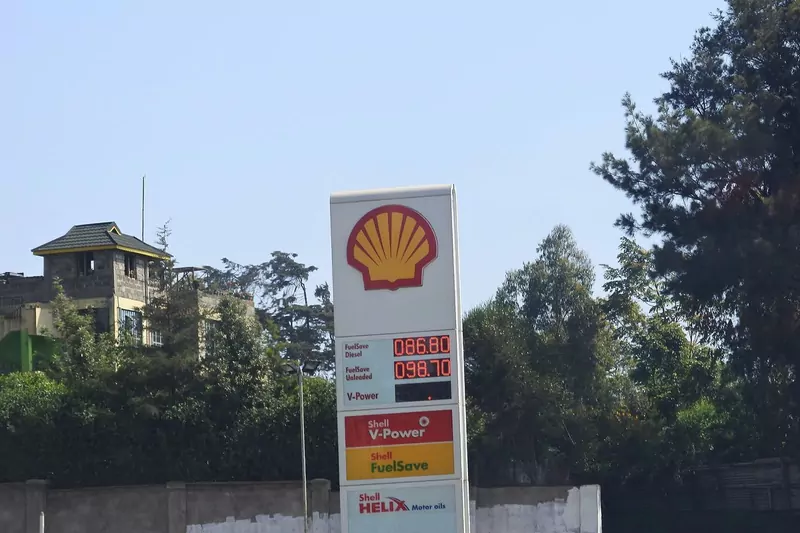A tall white Shell sign with the yellow and red shell logo and a digital price board, with trees and a house behind it under a blue sky