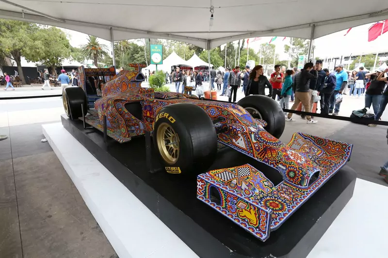 A Formula 1 car covered in colourful beadwork on display under a white tent, with visitors walking past