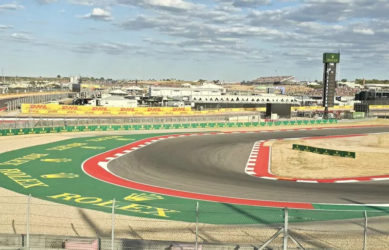 The Circuit of the Americas track at turn 15 under a blue sky, with grandstands and the Rolex timing tower in the distance