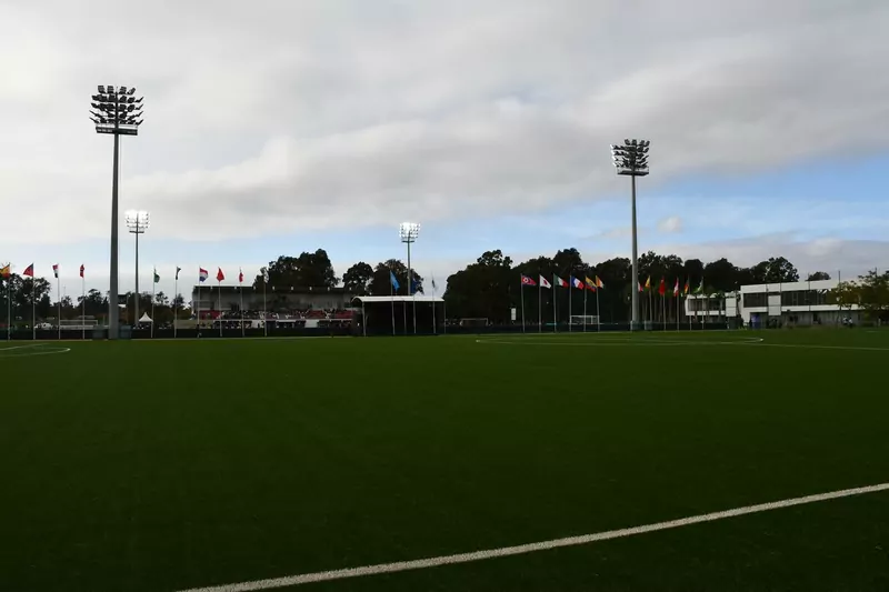 A dark green pitch seen from the touchline under floodlights, with a row of national flags and a small stand in the distance beneath cloudy skies