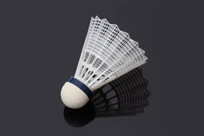 A white nylon badminton shuttlecock with a navy band above its rounded base, lying on a glossy dark surface with its shadow beside it