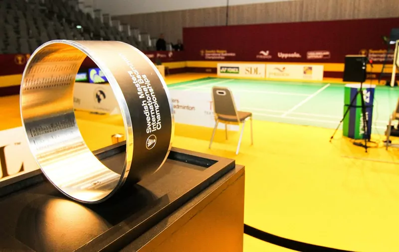 A silver ring-shaped trophy on a black stand in the foreground, with an empty green badminton court, a single chair and Yonex advertising boards behind it