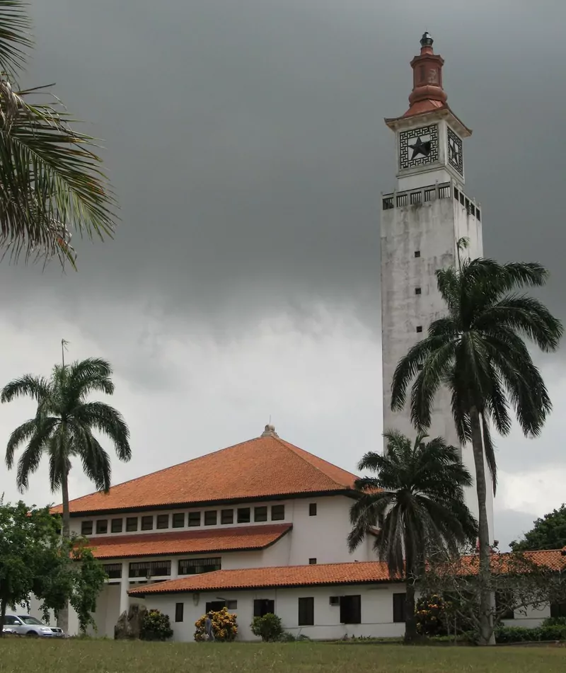 The white clock tower and red-tiled halls of the University of Ghana campus at Legon, Accra, under a dark rain cloud, with palm trees in front