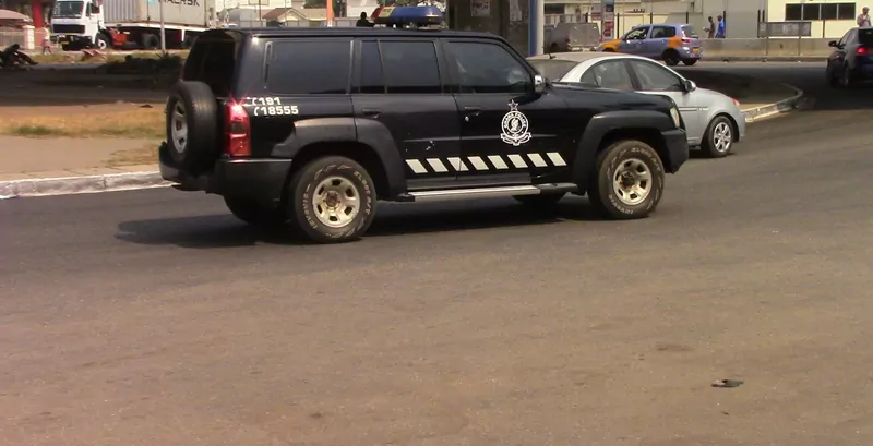 A black Ghana Police Service patrol SUV with the police crest on the door and a blue light on the roof drives through a junction in Ghana, with a silver car and a flyover pillar behind it