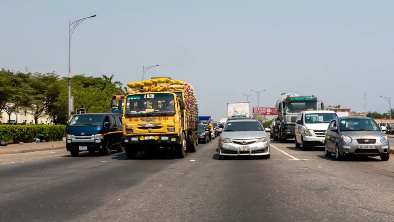 Cars, minibuses and a yellow lorry loaded with sacks wait side by side on a wide road in Accra, with a concrete mixer truck behind them and Ghanaian number plates on the vehicles