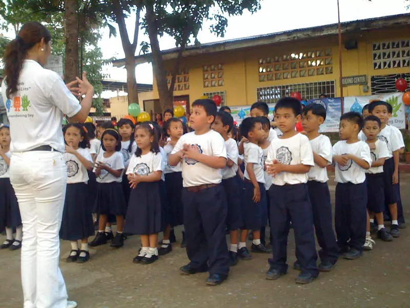 Schoolchildren in white shirts and navy uniforms copying a teacher's handwashing movements in a school yard