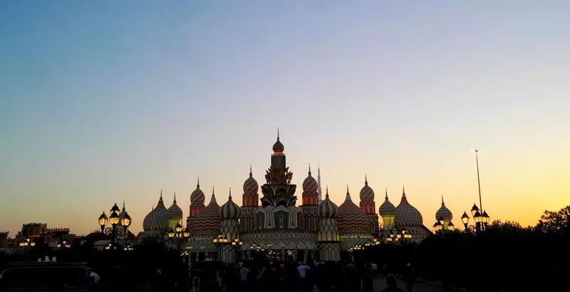 Floodlit domes and spires of the main entrance building at Global Village in Dubai, silhouetted against a dusk sky that fades from blue to orange, with lamp posts and a crowd below