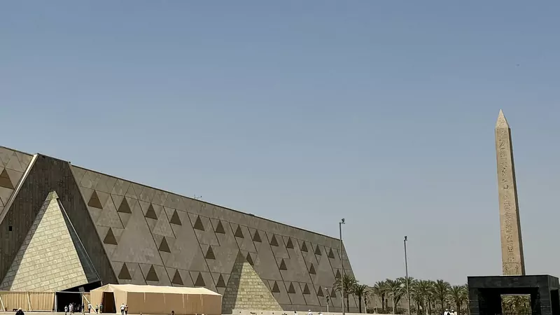 The long sloping stone wall of the Grand Egyptian Museum with a glass pyramid entrance, and a tall obelisk beside a black gate on the wide plaza, under a clear blue sky