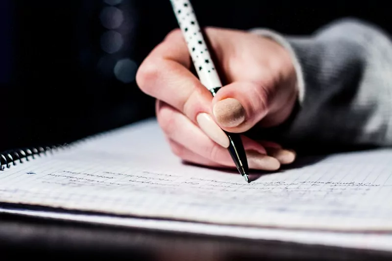 A hand holding a white pen with black dots and writing in a spiral notebook on a dark desk