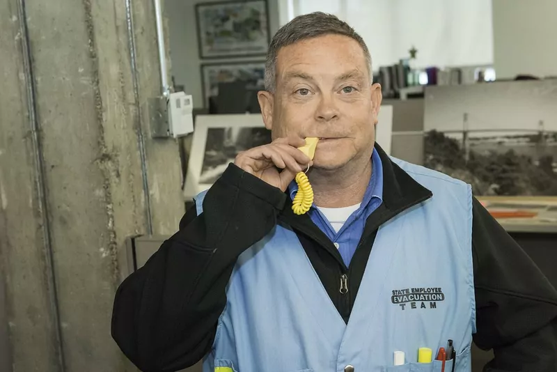 A man in a light blue evacuation team vest blowing a yellow whistle in an office to start an earthquake drill